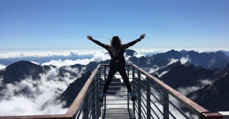 Unconventional Success - Person Standing on Hand Rails With Arms Wide Open Facing the Mountains and Clouds