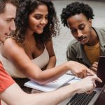 University Startups - Positive multiethnic male and female students sitting together with African American friend in cafeteria and showing with finger project details on laptop