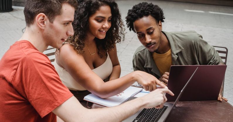 University Startups - Positive multiethnic male and female students sitting together with African American friend in cafeteria and showing with finger project details on laptop
