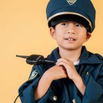 Career Goals - Pleasant Asian boy in police uniform and cap looking away while standing with hands near chest in studio on yellow background