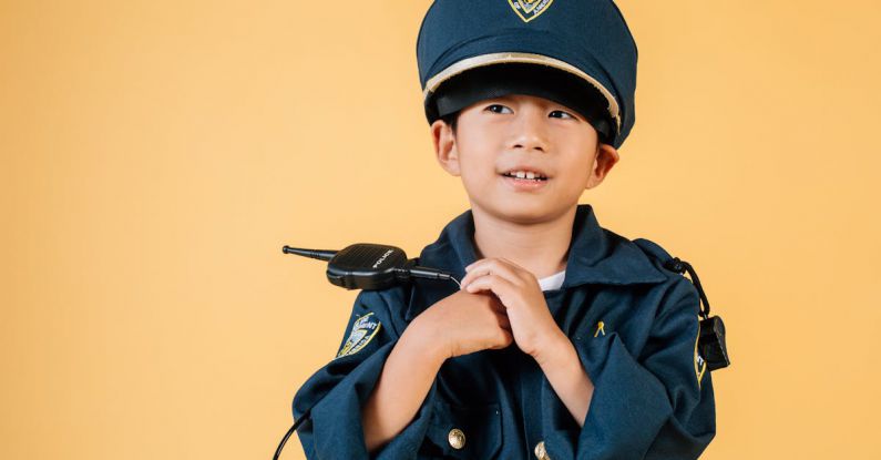 Career Goals - Pleasant Asian boy in police uniform and cap looking away while standing with hands near chest in studio on yellow background