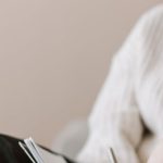 Task Delegation - Attentive female wearing eyeglasses and casual outfit sitting barefoot with crossed legs on comfortable couch in modern flat and taking notes in notebook with pen