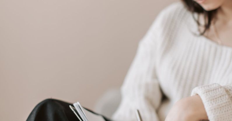 Task Delegation - Attentive female wearing eyeglasses and casual outfit sitting barefoot with crossed legs on comfortable couch in modern flat and taking notes in notebook with pen